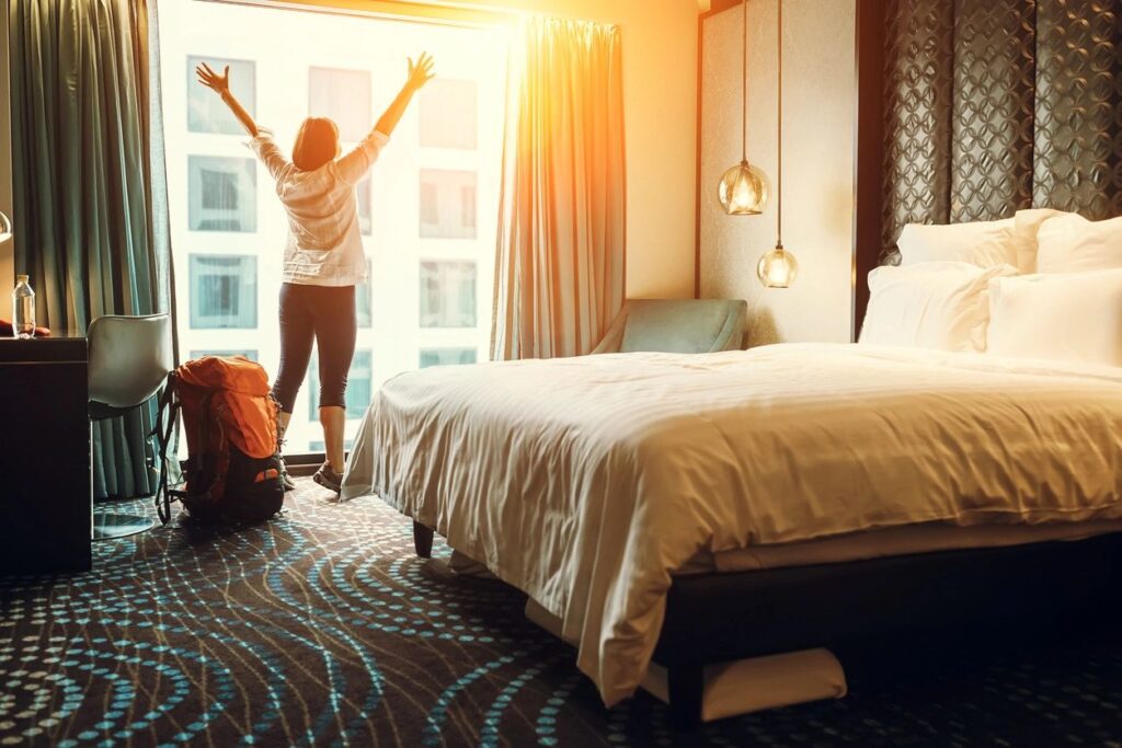 a picture of a woman with her arms extended upwards making a "Y" shape. She is standing in front of a window next to a big bed that's made, and a red luggage bag next to her